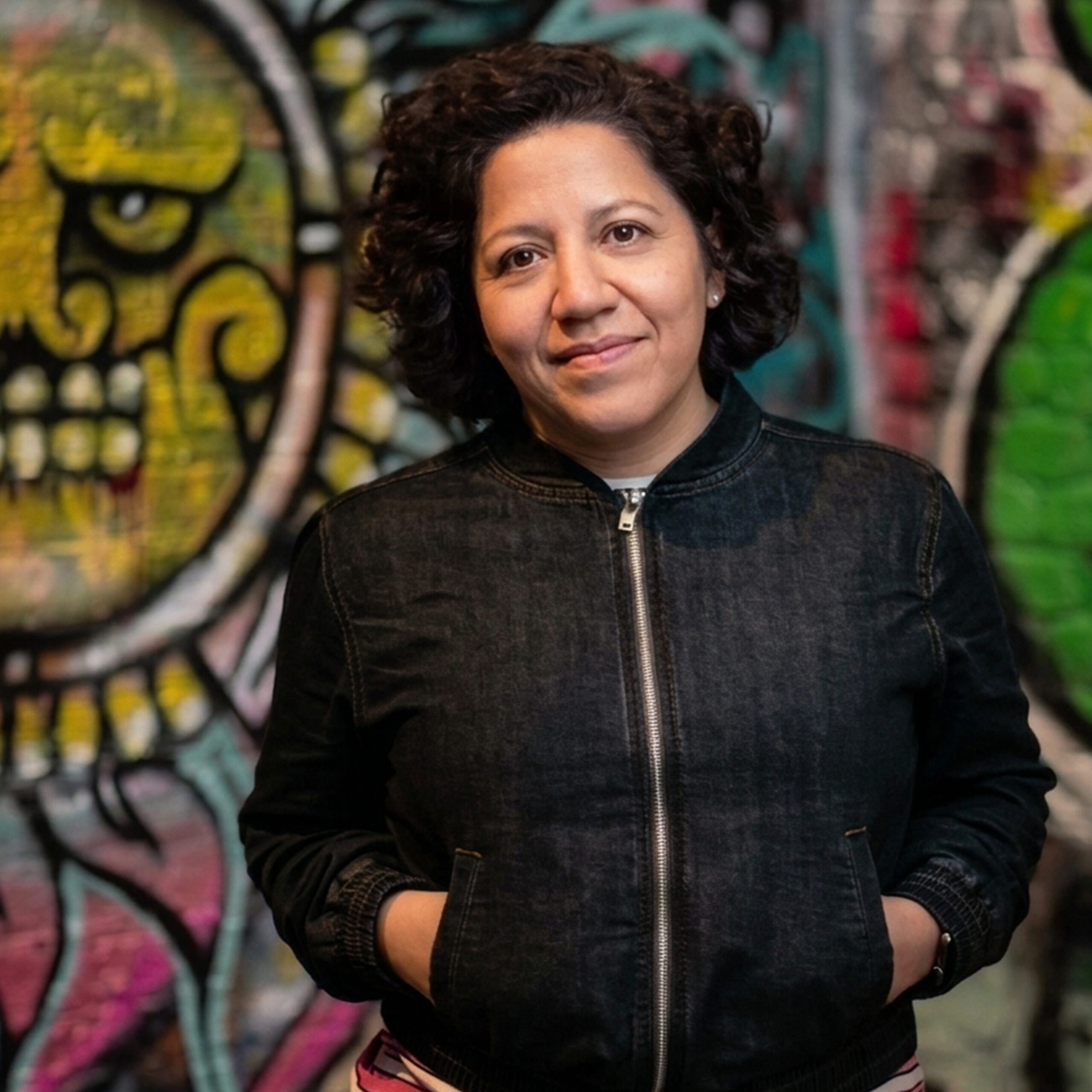 Porträt von Teresa, eine Frau mit lockigem dunklem Haar und einem leichten Lächeln, in einer dunklen Jeansjacke, vor einer bunten Graffiti-Wand
