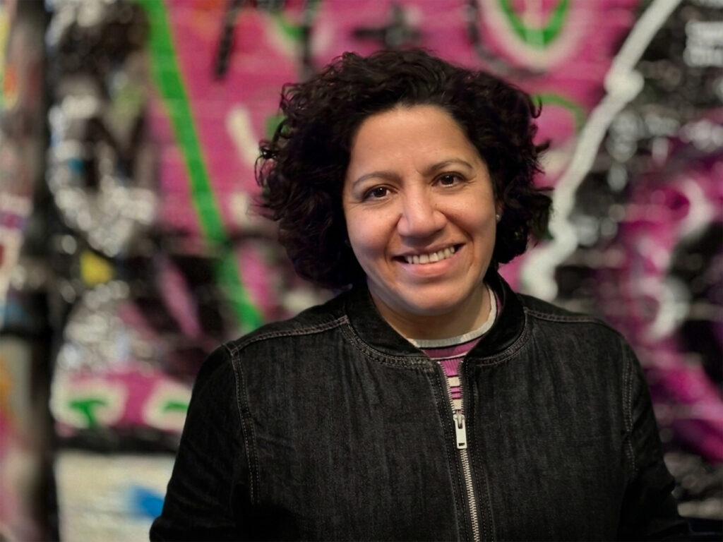 Porträtfoto einer lächelnden Frau mit dunklen lockigen Haaren, in einer schwarzen Jeansjacke, vor einem bunten Graffiti-Hintergrund in Pink, Grün und Weiß."